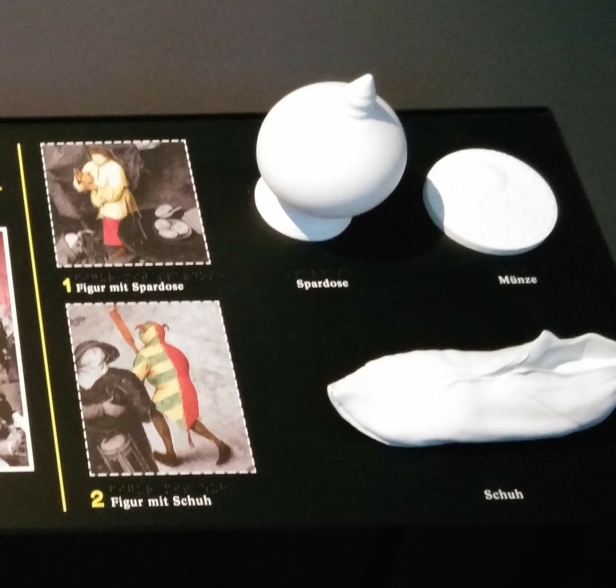 Zu sehen sind Details von einem Bild mit Tastgegenständen: eine Spardose, ein mittelalterlicher Schuh und eine Münze
